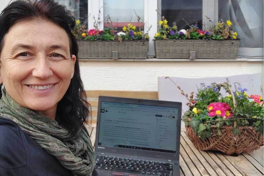 Marianne sitzt am Gartentisch im Innenhof umringt von bunten Frühlingsblühern auf dem Tisch und am Fenster. Hier lässt sich die transformierende, zentrierende Kraft des Schreibens spüren.