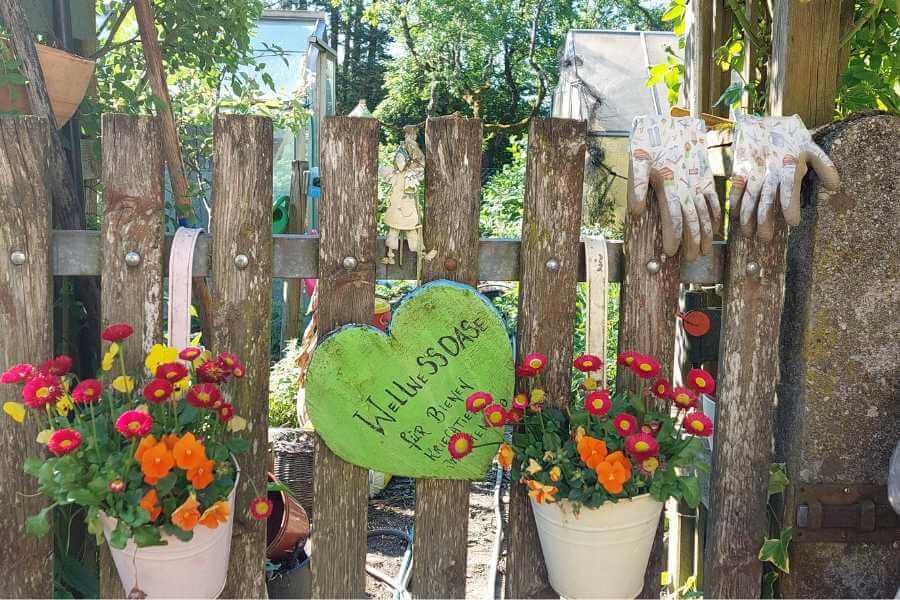 Die Gartenpforte zum Bauerngarten mit Schild ein Holzherz mit Aufschrift: Wllnessoase für Bienen, Kriechtiere und Insekten. Es hängen Gartenhandschuhe daran und zwei bunte Blumentöpfe