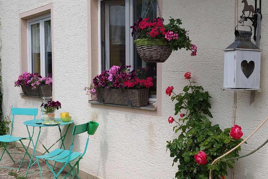 Ein kleiner Sitzplatz zwei gründe Metallstühle und Tisch mit Blumen und auch an den Fensterns des Hausen herrliche lila-pink-rosa Blumen. Am Hauseingang herrliche englische Rosen und Blumenampel. Eine Hängelaterne aus Holz in weiß mit Herz. 