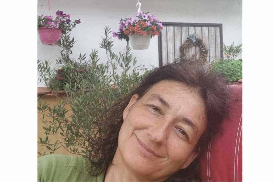 Marianne angelehnt im Gartenstuhl auf der Terrasse im Hintergrund Blumenampeln. Sie lächelt.